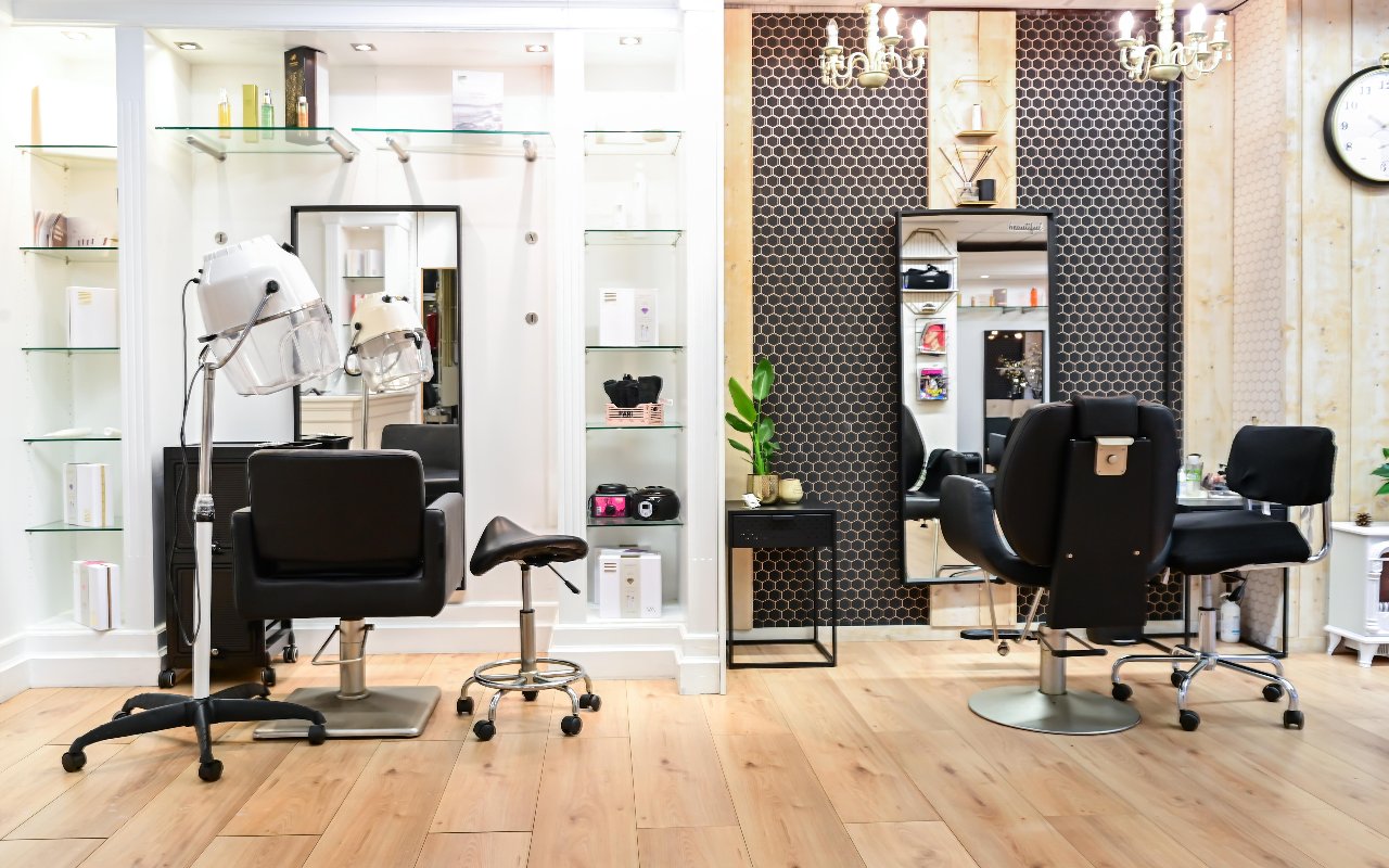 De kapsalon: twee kappersstoelen voor spiegels met glazen planken, een droogkap en een zwarte tegelwand